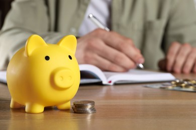 Photo of Man at wooden table, focus on yellow piggy bank. Space for text