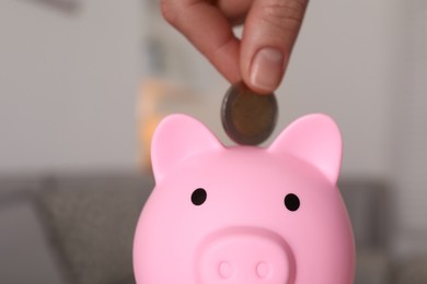 Woman putting coin into pink piggy bank on blurred background, closeup