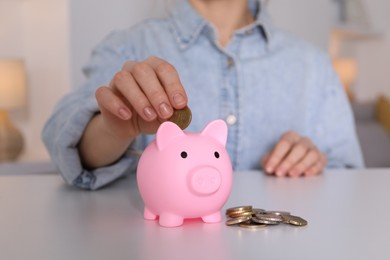 Woman putting coin into pink piggy bank at white table, closeup