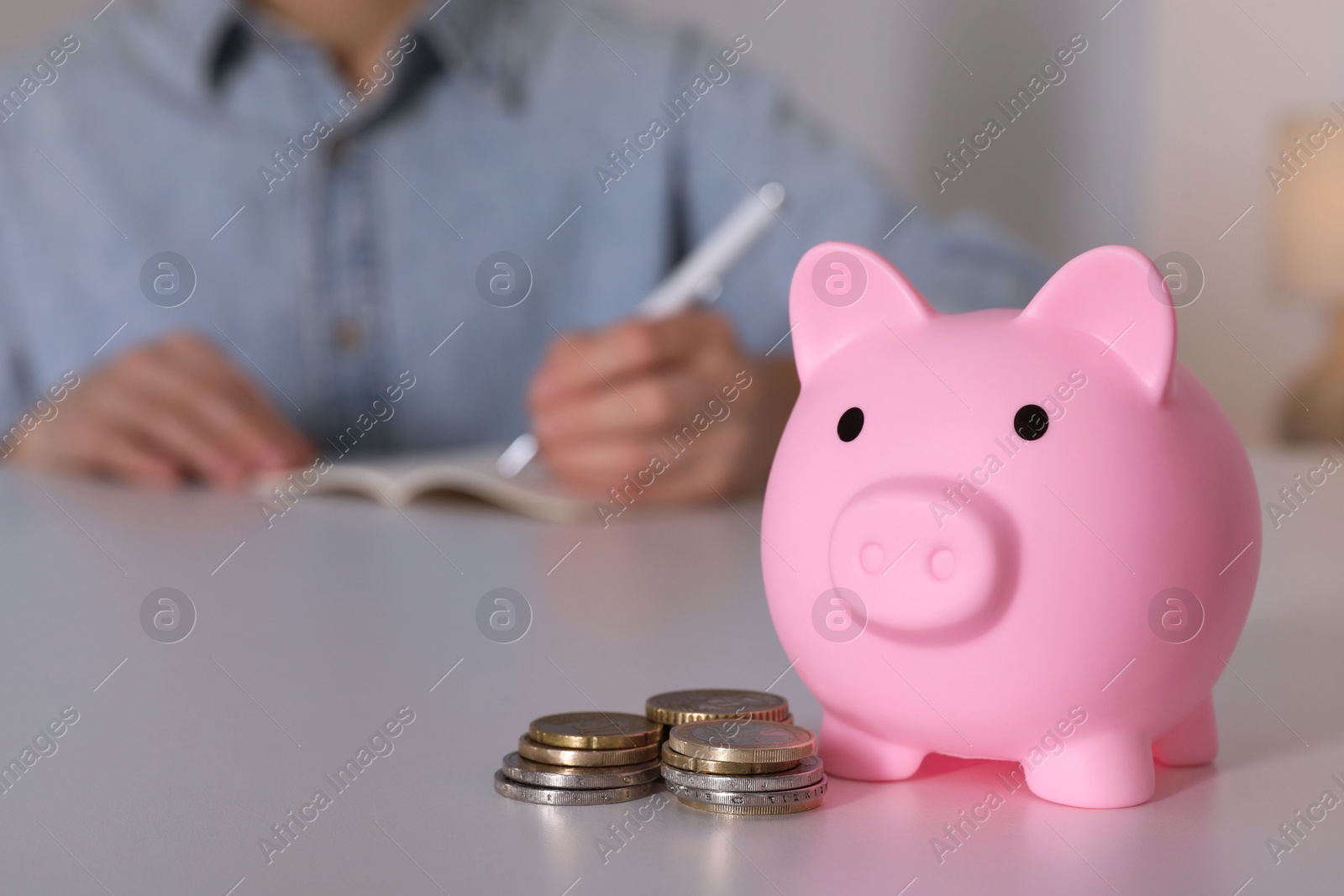 Photo of Woman at white table, focus on pink piggy bank. Space for text