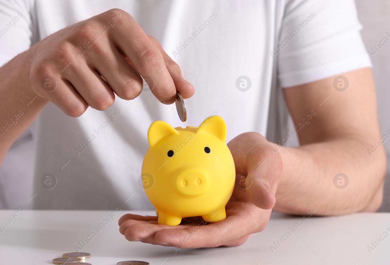 Photo of Man putting coin into yellow piggy bank at white table, closeup