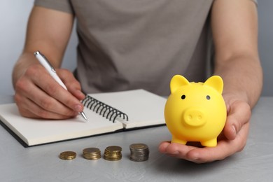 Photo of Man with yellow piggy bank, coins and notebook at grey table, closeup