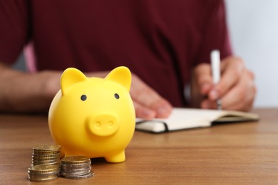 Photo of Man at wooden table, focus on yellow piggy bank and coins. Space for text