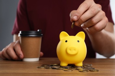 Photo of Man putting coin into yellow piggy bank at wooden table, closeup