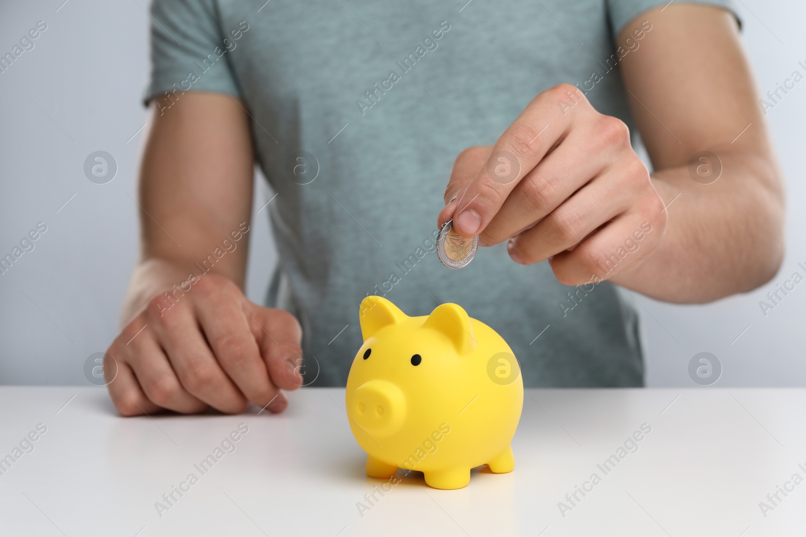 Photo of Man putting coin into yellow piggy bank at white table, closeup