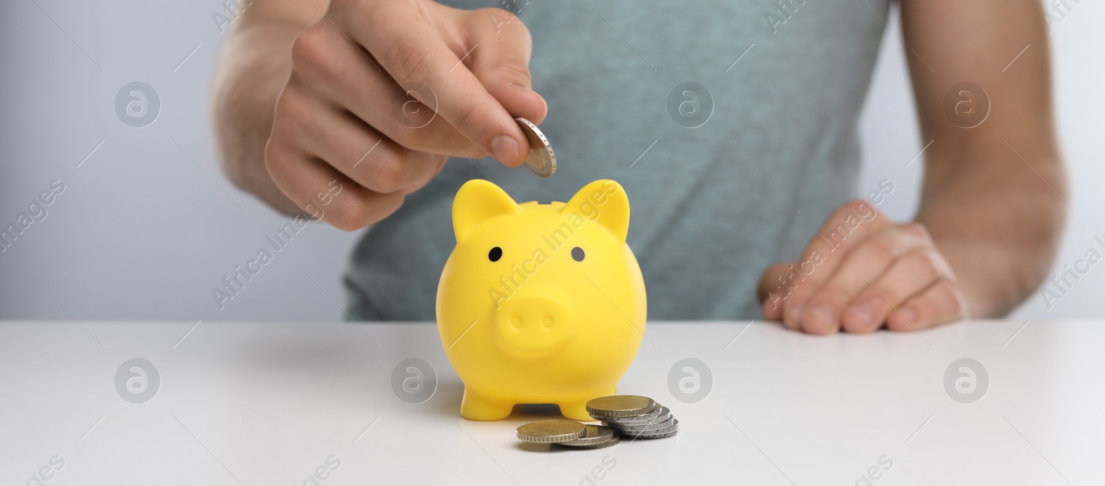 Photo of Man putting coin into yellow piggy bank at white table, closeup