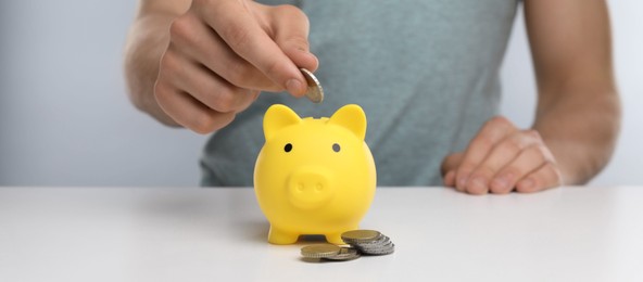 Photo of Man putting coin into yellow piggy bank at white table, closeup