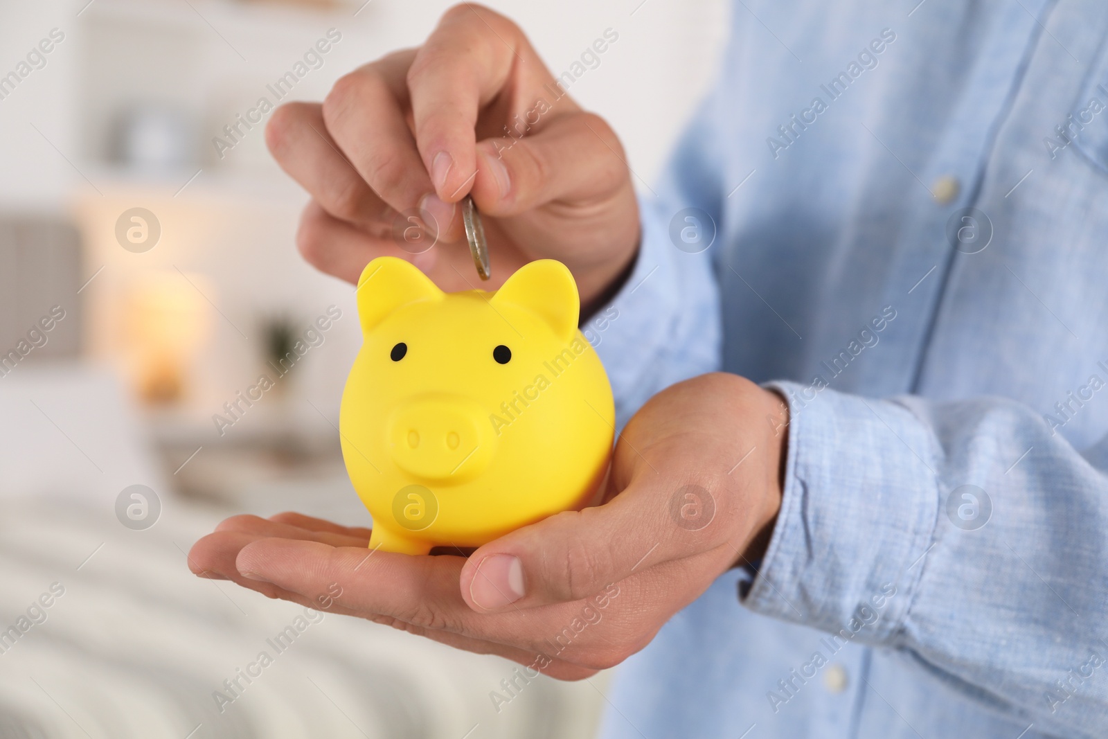 Photo of Man putting coin into yellow piggy bank indoors, closeup