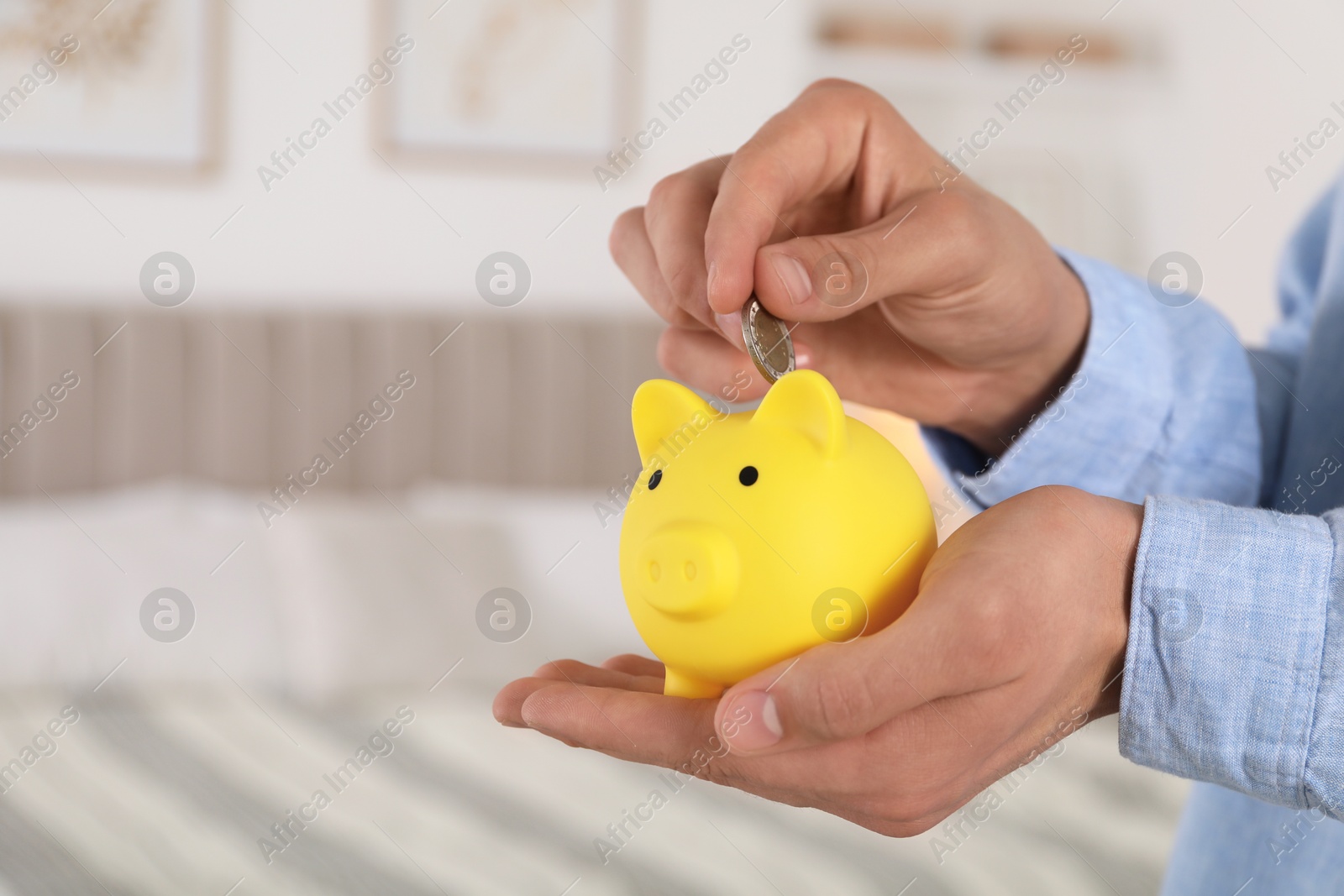 Photo of Man putting coin into yellow piggy bank indoors, closeup. Space for text