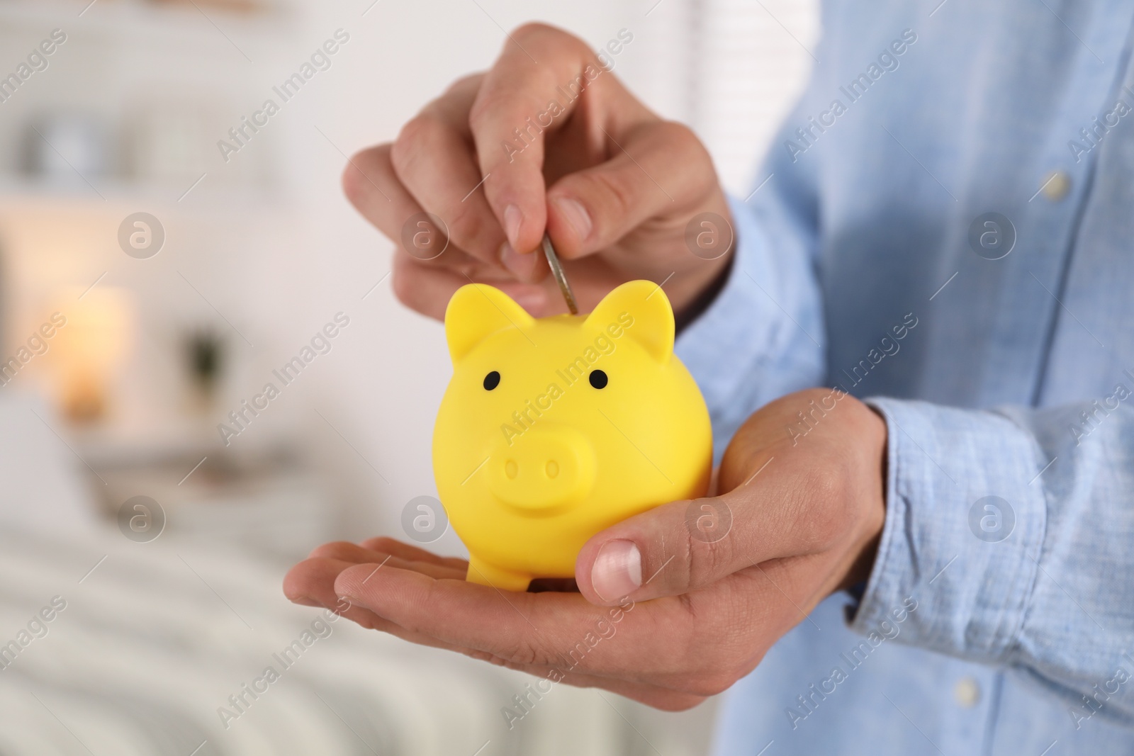 Photo of Man putting coin into yellow piggy bank indoors, closeup