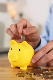 Photo of Man putting coin into yellow piggy bank on wooden table, closeup