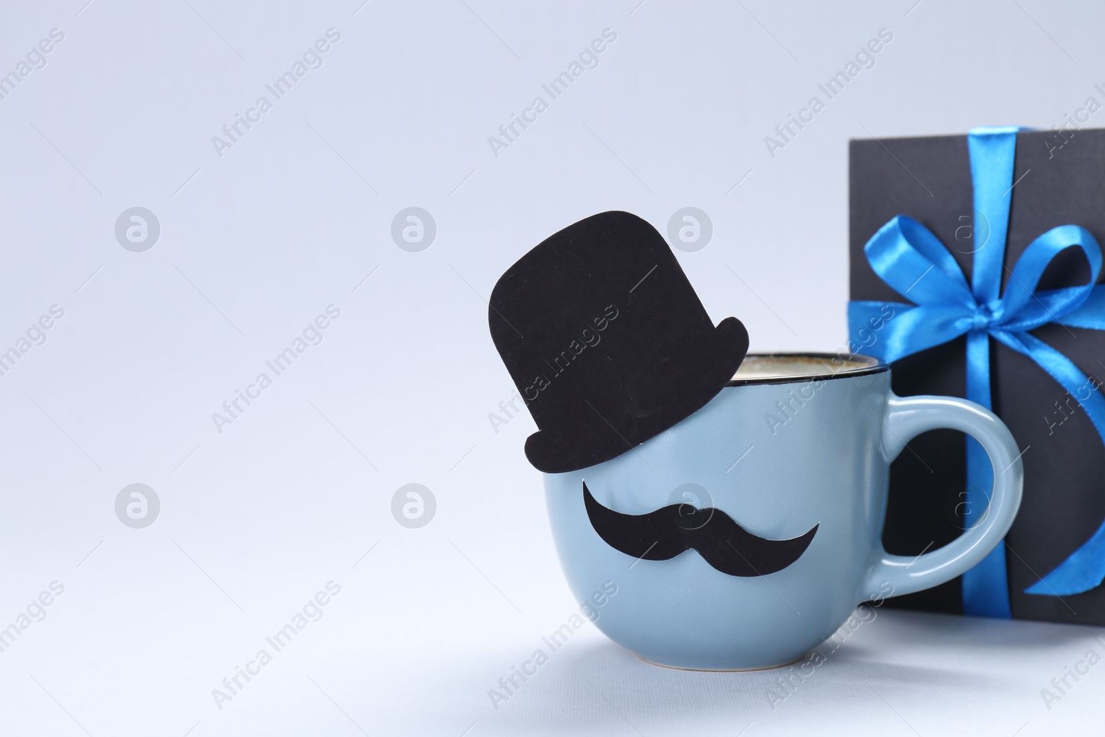 Photo of Happy Father's Day. Cup of coffee with funny paper hat, moustache and gift on light background. Space for text