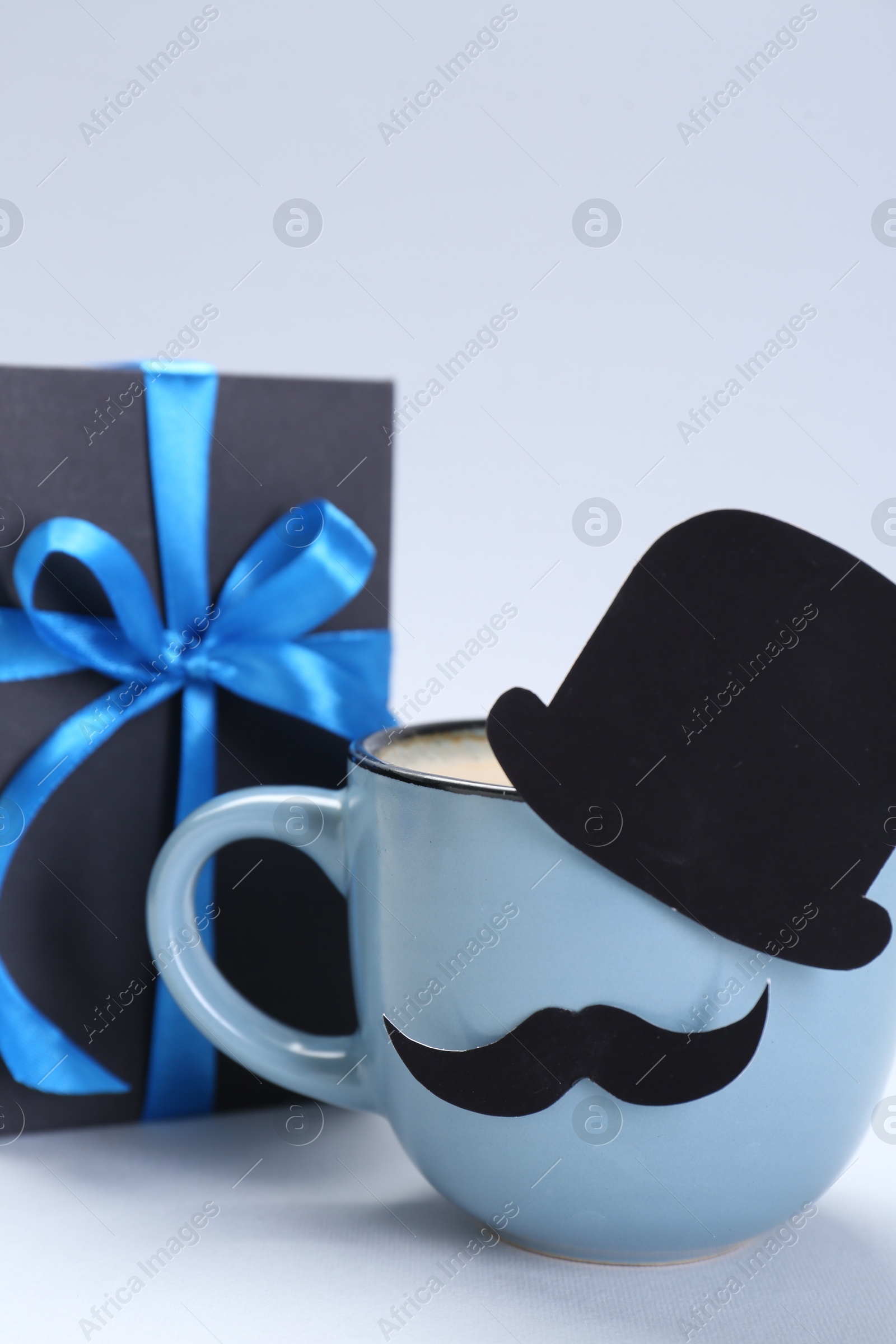 Photo of Happy Father's Day. Cup of coffee with funny paper hat, moustache and gift on light background