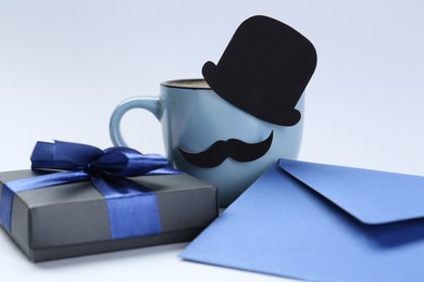 Photo of Happy Father's Day. Cup of coffee with funny paper hat, moustache, gift and envelope on light background