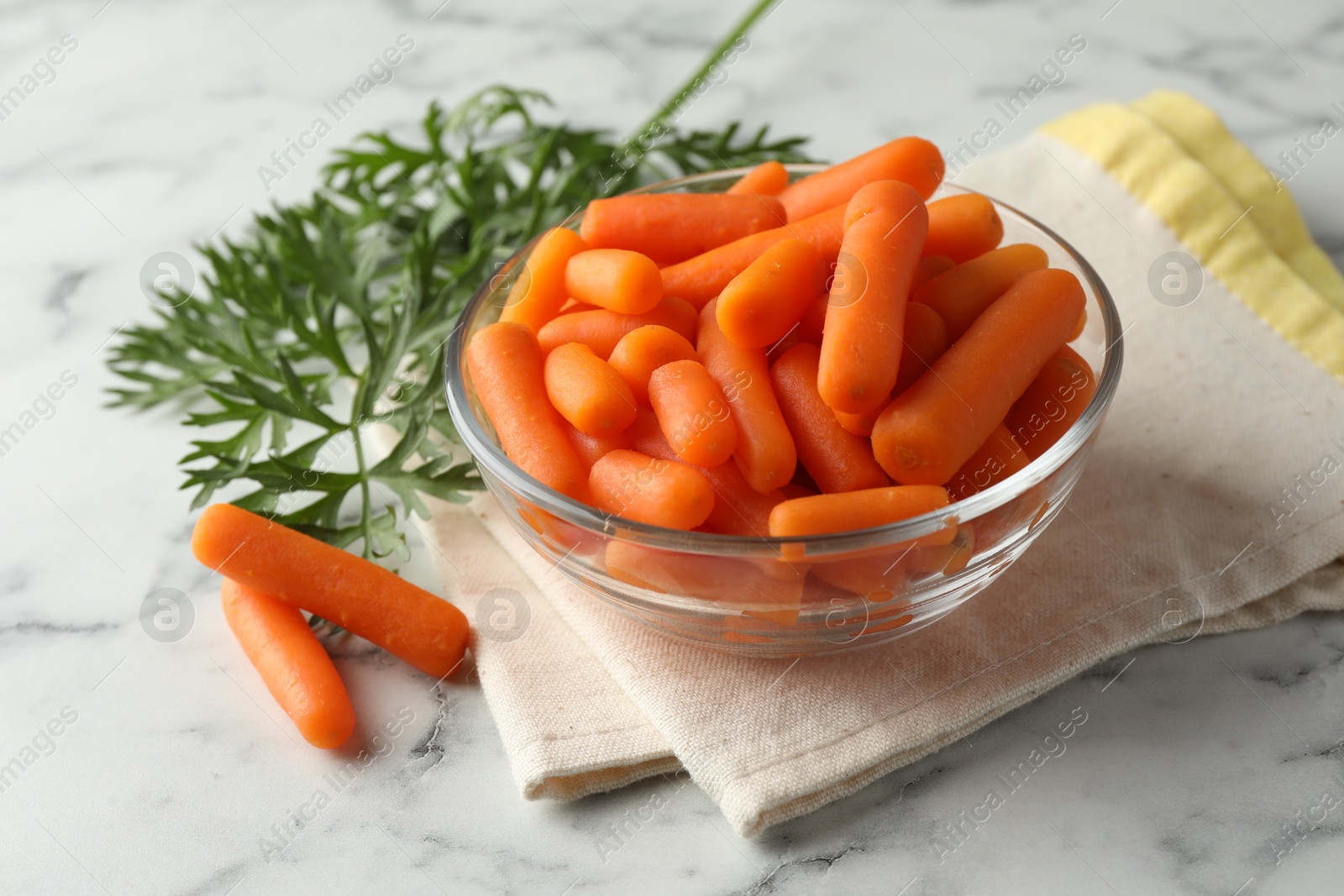 Photo of Baby carrots in bowl and green leaves on white marble table