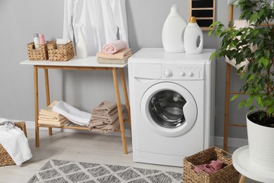 Photo of Washing machine, storage bench, detergents, laundry and houseplant indoors