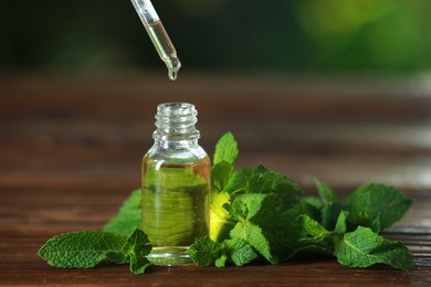 Photo of Dripping mint essential oil from pipette into bottle of on wooden table, closeup