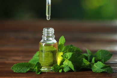 Photo of Dripping mint essential oil from pipette into bottle of on wooden table, closeup