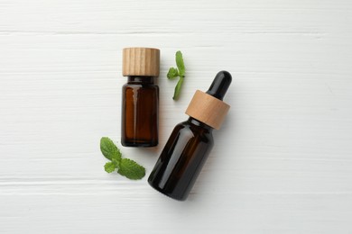 Bottles of mint essential oil and fresh leaves on white wooden table, top view