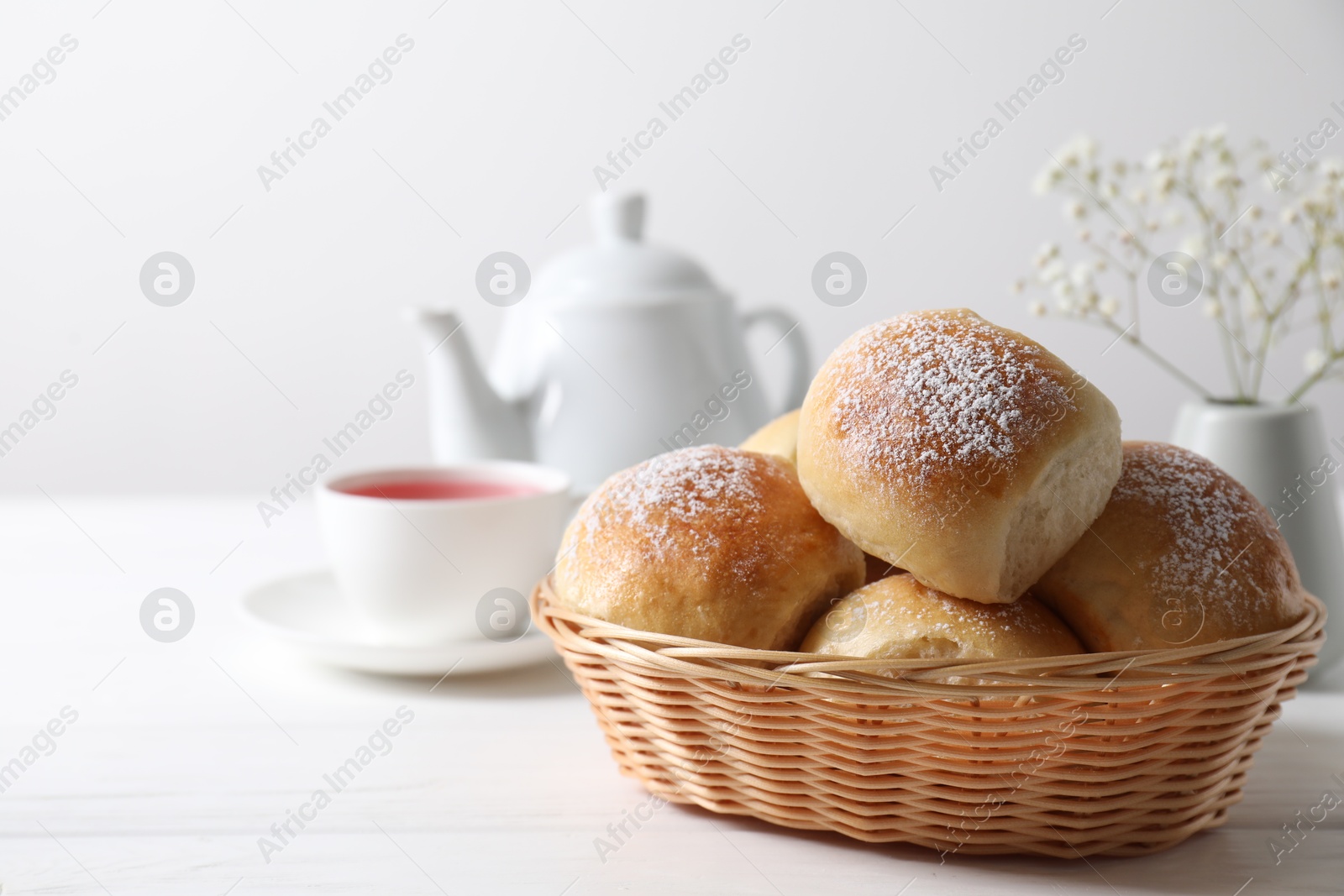 Photo of Delicious dough balls with powdered sugar in basket, tea, teapot and gypsophila flowers on white wooden table