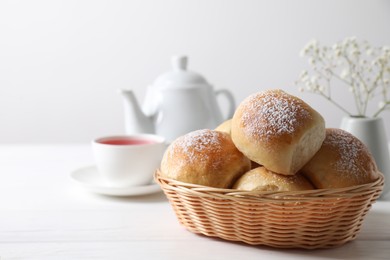 Photo of Delicious dough balls with powdered sugar in basket, tea, teapot and gypsophila flowers on white wooden table