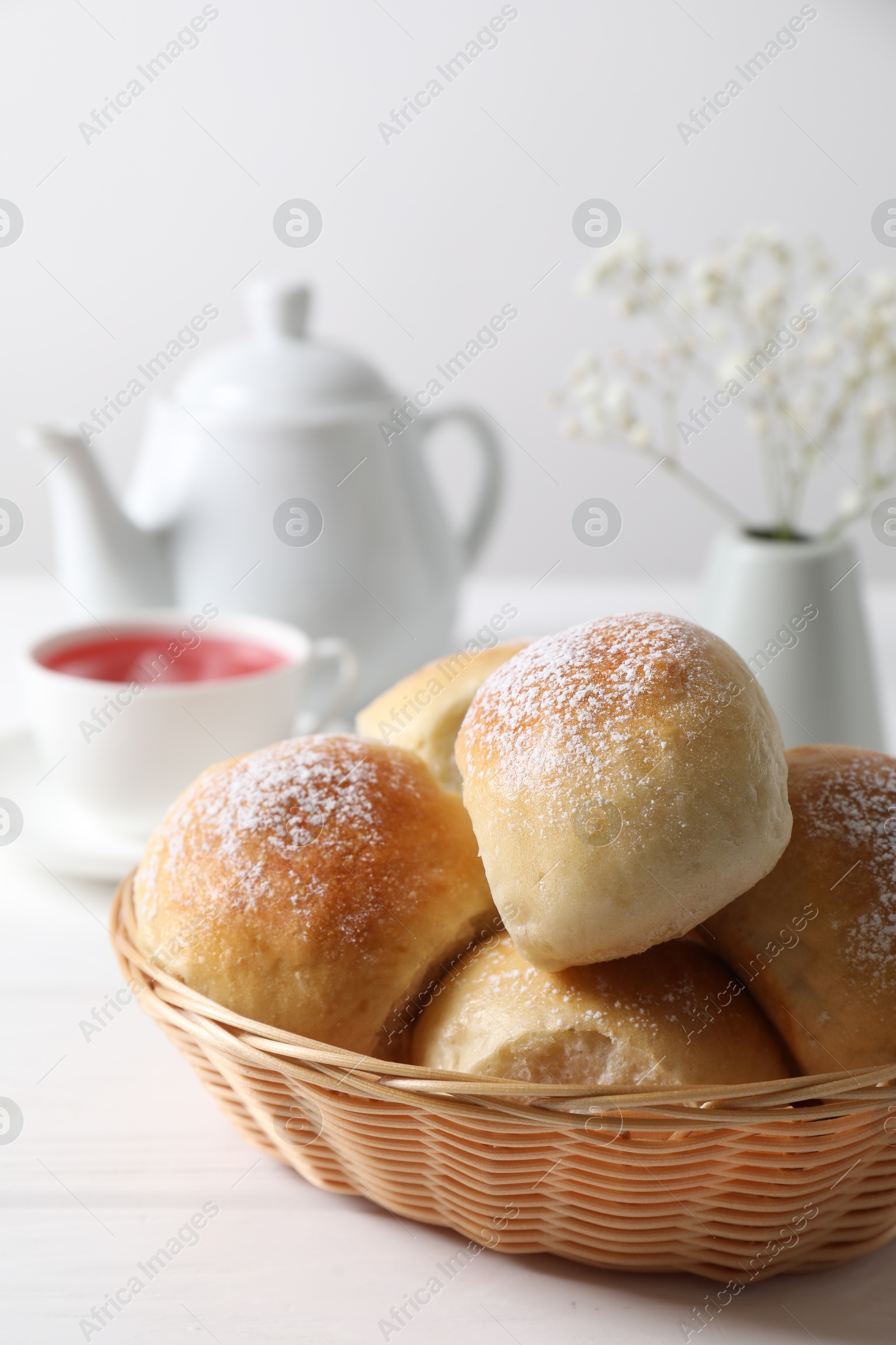 Photo of Delicious dough balls with powdered sugar in basket, tea, teapot and gypsophila flowers on white wooden table