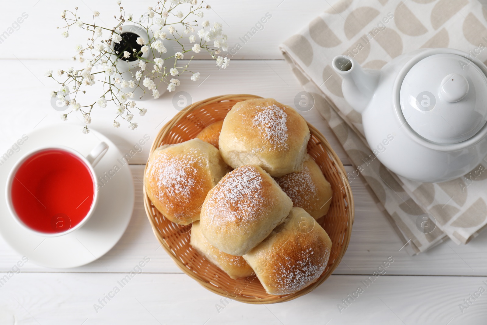 Photo of Delicious dough balls with powdered sugar in basket, tea, teapot and gypsophila flowers on white wooden table, top view