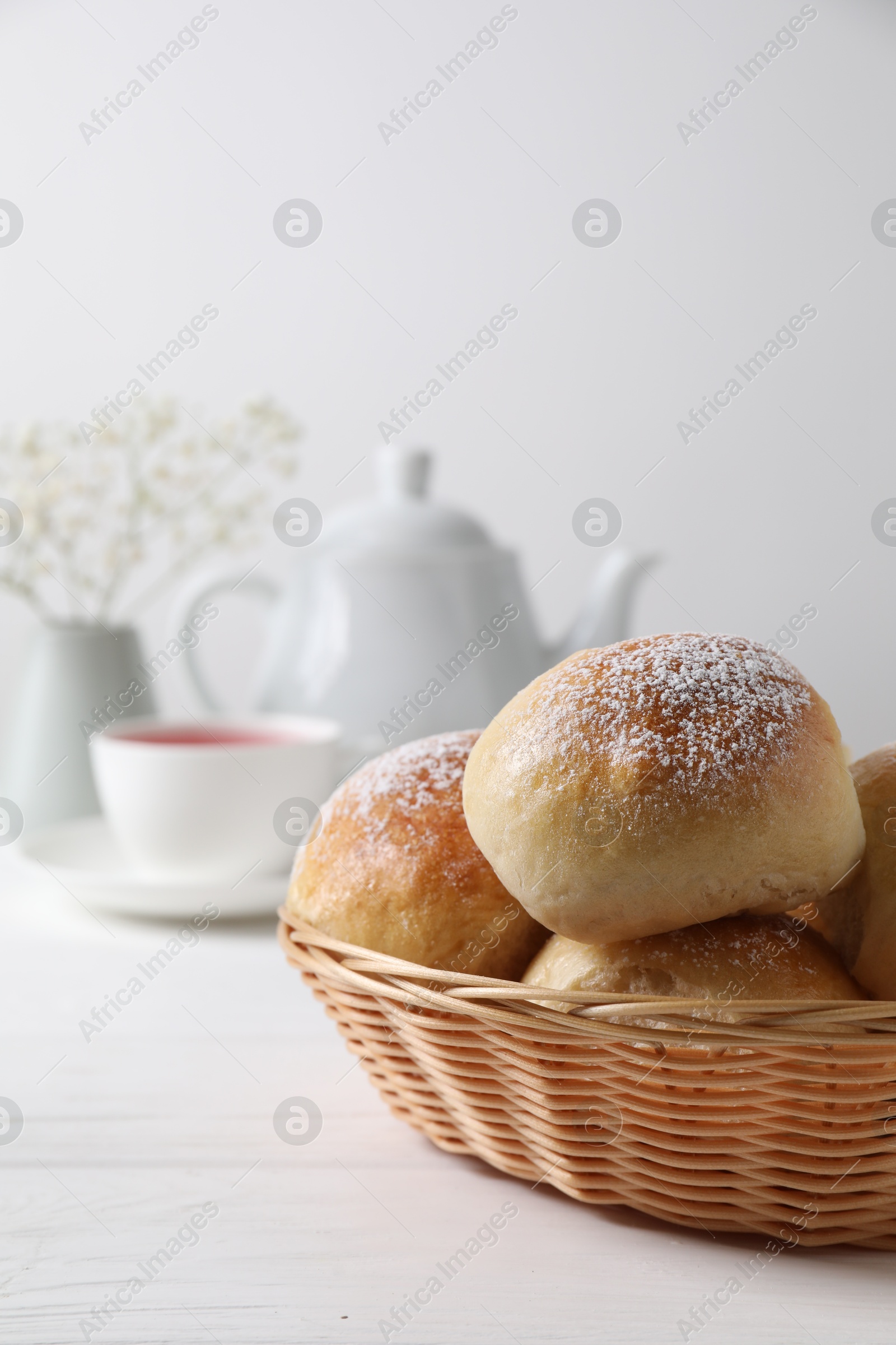 Photo of Delicious dough balls with powdered sugar in basket, tea, teapot and gypsophila flowers on white wooden table