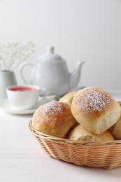 Photo of Delicious dough balls with powdered sugar in basket, tea, teapot and gypsophila flowers on white wooden table