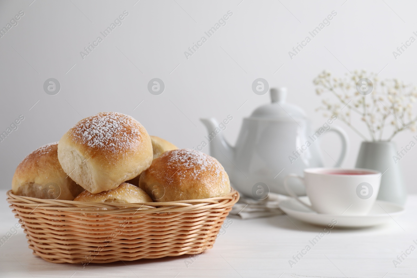 Photo of Delicious dough balls with powdered sugar in basket, tea, teapot and gypsophila flowers on white wooden table