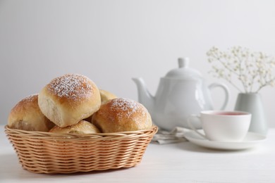 Photo of Delicious dough balls with powdered sugar in basket, tea, teapot and gypsophila flowers on white wooden table