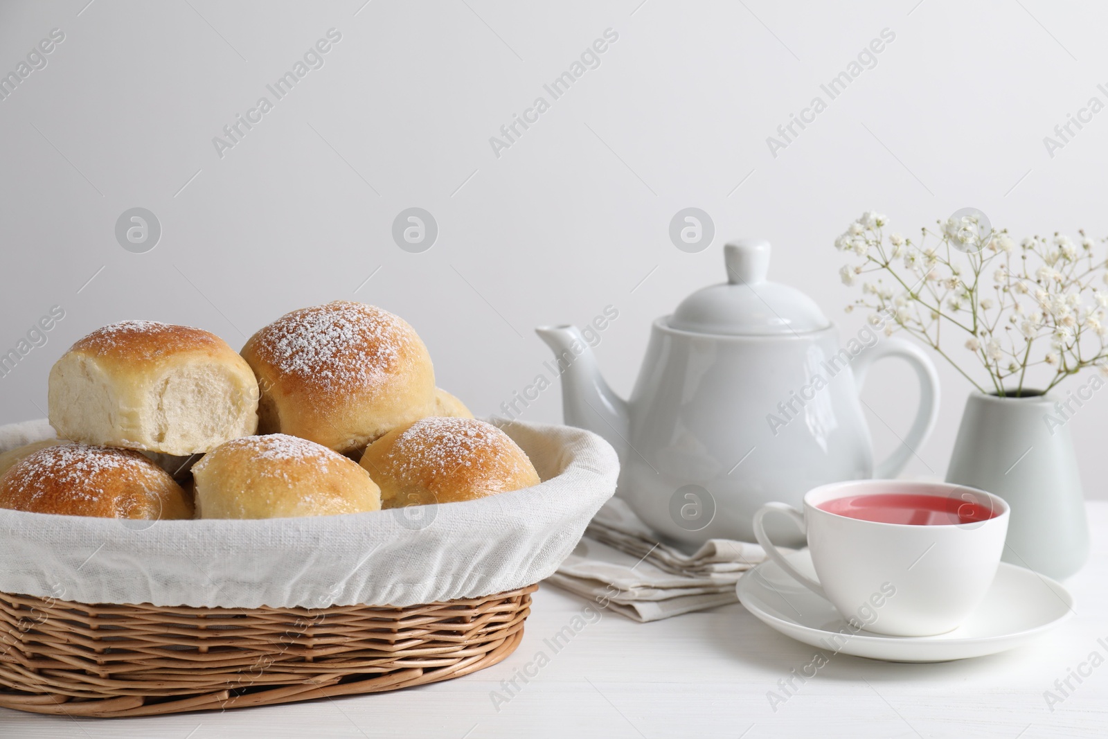 Photo of Delicious dough balls with powdered sugar in basket, tea, teapot and gypsophila flowers on white wooden table