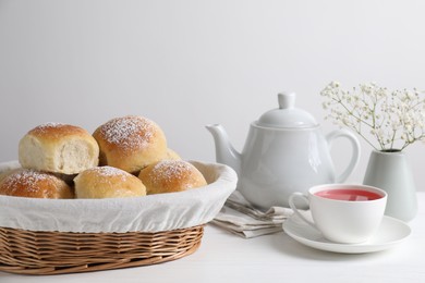 Photo of Delicious dough balls with powdered sugar in basket, tea, teapot and gypsophila flowers on white wooden table