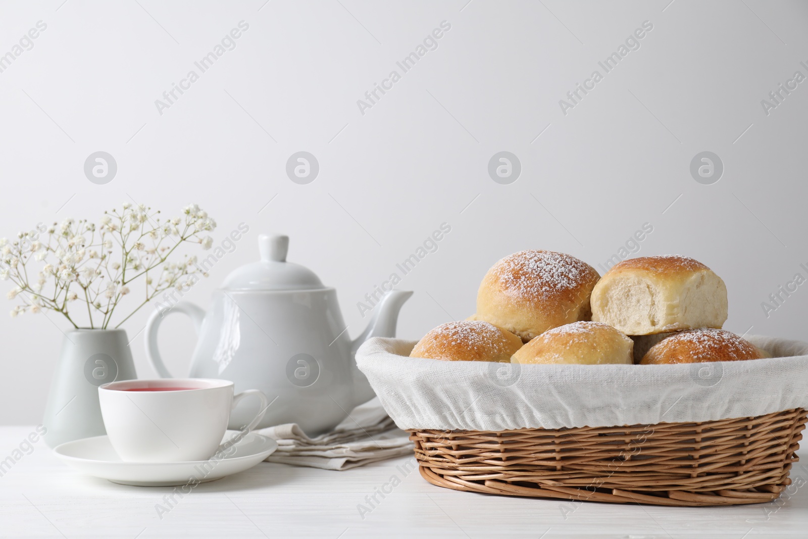 Photo of Delicious dough balls with powdered sugar in basket, tea, teapot and gypsophila flowers on white wooden table