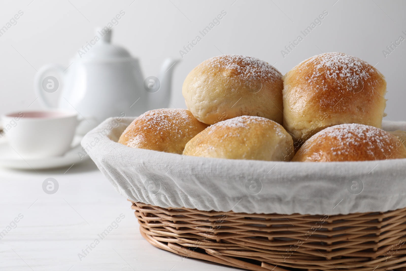 Photo of Delicious dough balls with powdered sugar in basket, tea and teapot on white wooden table, closeup