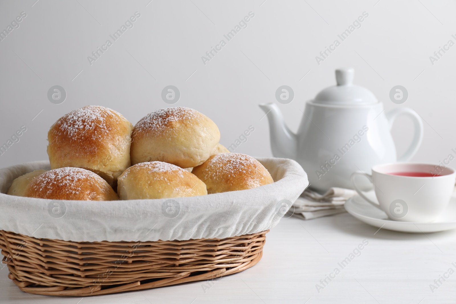 Photo of Delicious dough balls with powdered sugar in basket, tea and teapot on white wooden table