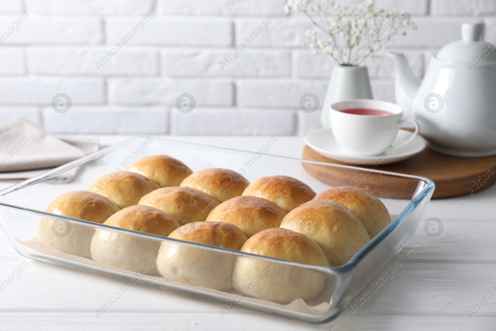 Photo of Delicious dough balls in baking dish, tea, teapot and gypsophila flowers on white wooden table