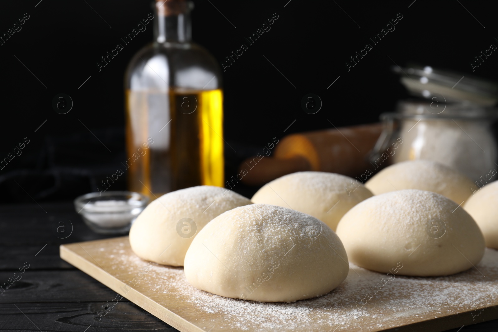 Photo of Raw dough balls, oil, flour and rolling pin on black wooden table