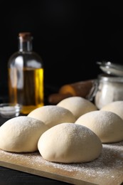 Photo of Raw dough balls, oil, flour and rolling pin on black wooden table