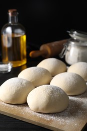 Photo of Raw dough balls, oil, flour and rolling pin on black wooden table