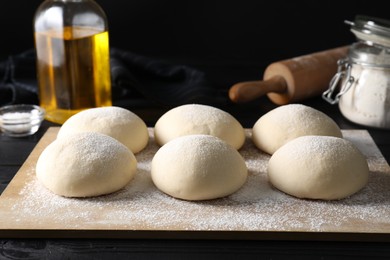 Photo of Raw dough balls, oil, flour and rolling pin on black wooden table