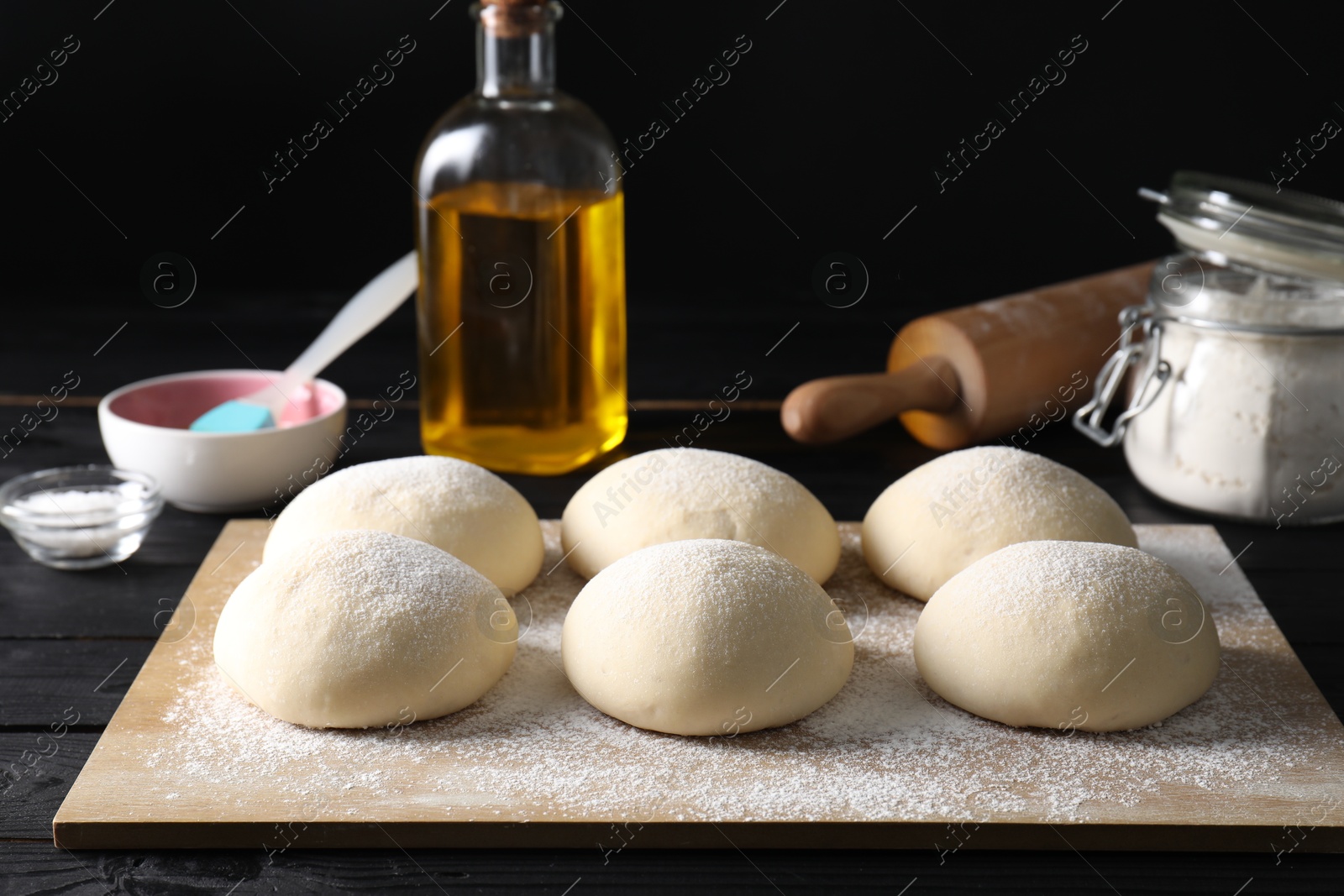 Photo of Raw dough balls, oil, flour, salt and rolling pin on black wooden table