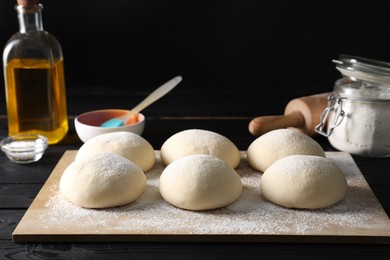 Photo of Raw dough balls, oil, flour, salt and rolling pin on black wooden table