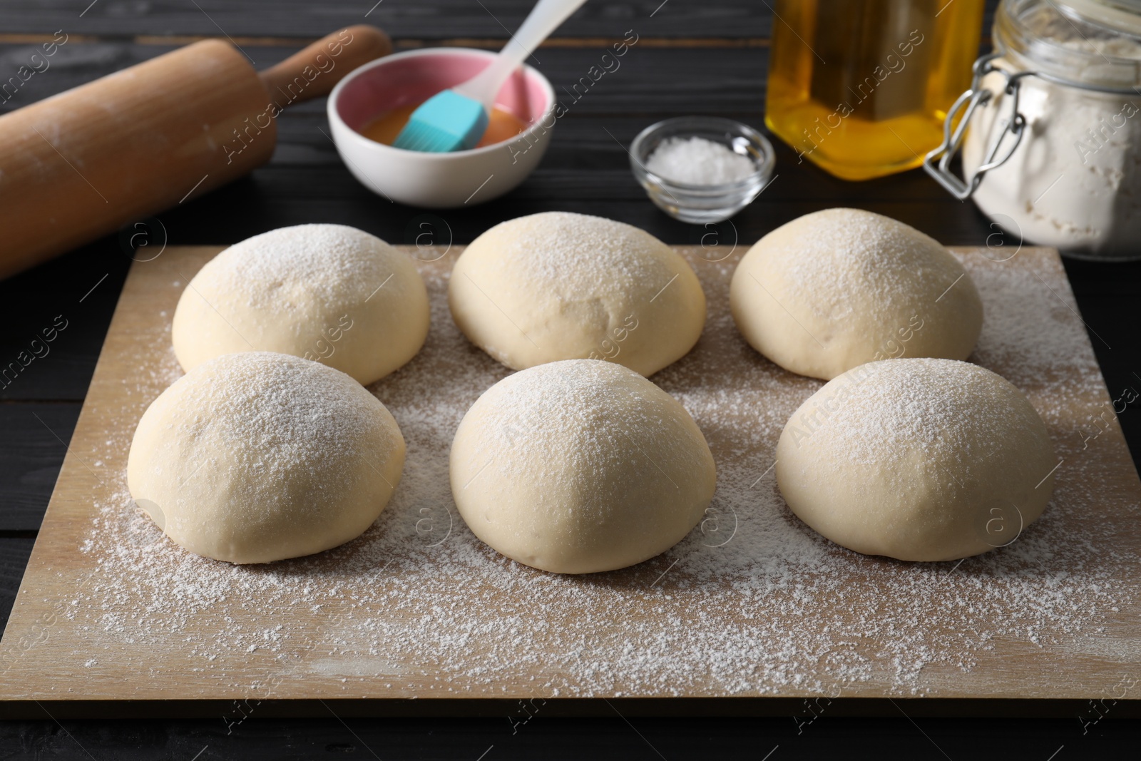 Photo of Raw dough balls, yolk, oil, flour, salt and rolling pin on black wooden table