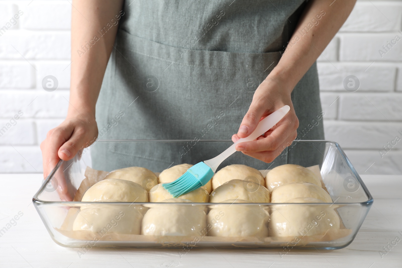 Photo of Woman spreading egg yolk onto raw dough balls at white wooden table, closeup