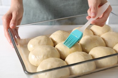 Woman spreading egg yolk onto raw dough balls at white wooden table, closeup