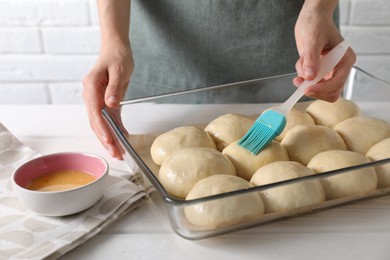 Photo of Woman spreading egg yolk onto raw dough balls at white wooden table, closeup