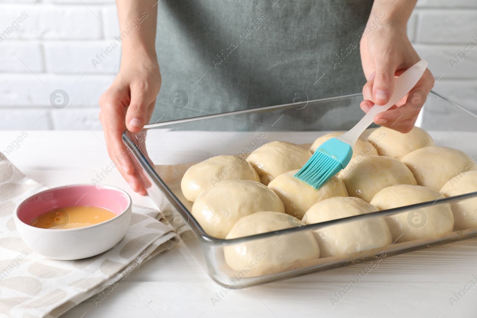 Photo of Woman spreading egg yolk onto raw dough balls at white wooden table, closeup