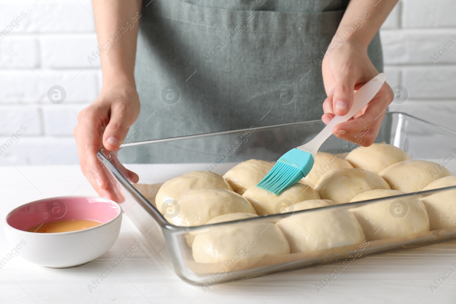 Photo of Woman spreading egg yolk onto raw dough balls at white wooden table, closeup