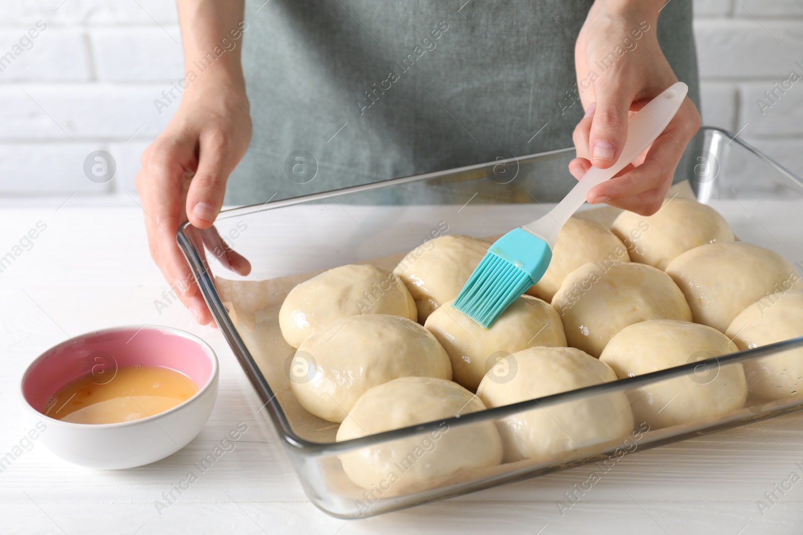 Photo of Woman spreading egg yolk onto raw dough balls at white wooden table, closeup